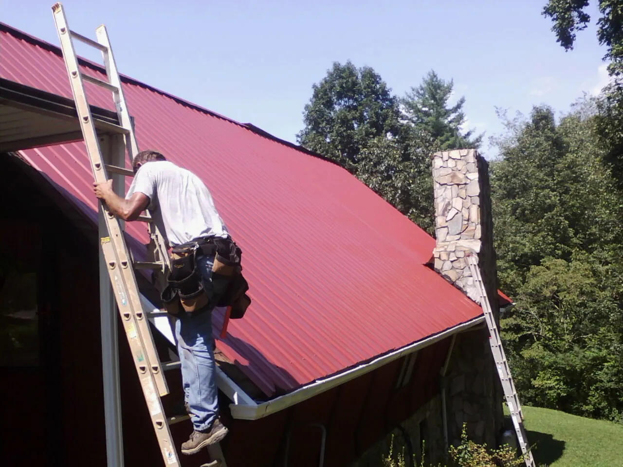 Roofer on a ladder beside a red metal roof and stone chimney — Soffit & Fascia Repair by Roofing Brothers in Culver